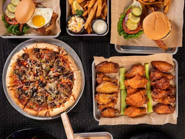 An overhead view of a black table with several different food items on it including: pizza, wings, a burger, and fries.